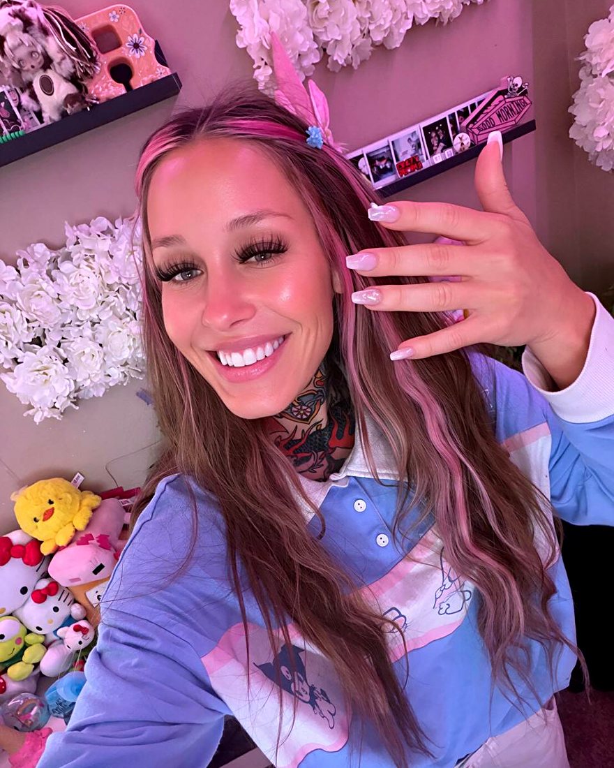 Amy with pink hair and long nails smiling in her pink room full of plushies